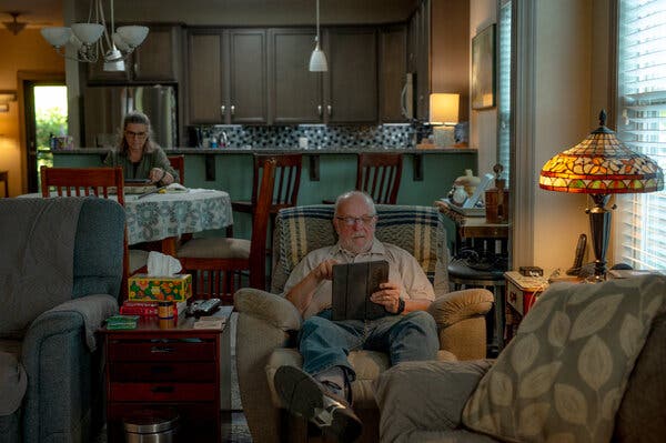 Mr. Wilczynski sitting in a reclining chair in his home, working on a computer. Ms. Brasier sits at the kitchen table at the rear.